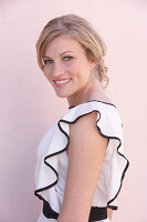 A blonde woman standing against a pink background wearing a light blouse with a black trim