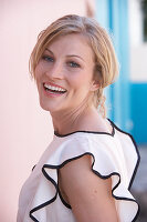 A blonde woman standing against a pink background wearing a light blouse with a black trim