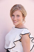 A blonde woman standing against a pink background wearing a light blouse with a black trim