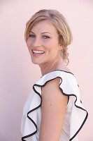 A blonde woman standing against a pink background wearing a light blouse with a black trim