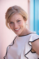 A blonde woman standing against a pink background wearing a light blouse with a black trim