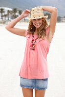 A young blonde woman on a beach wearing a pink top, a short denim skirt and a beige hat