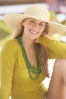 A young blonde woman on a beach wearing an olive top and a beige summer hat