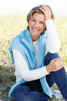 A mature woman with short blonde hair in the countryside wearing blue jeans and a white shirt with a blue jumper over her shoulders