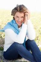A mature woman with short blonde hair in the countryside wearing blue jeans and a white shirt with a blue jumper over her shoulders