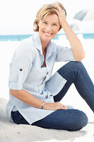A mature woman with short blonde hair on a beach wearing a light-blue striped short and blue jeans