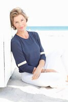 A mature woman with short blonde hair on a beach wearing a blue shirt and white trousers