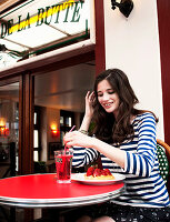 A young brunette woman wearing a striped shirt sitting in a cafe with a slice of cake and a drink