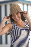 A young brunette woman on a beach wearing a blue-and-white striped top and a summer hat