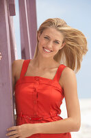 Junge blonde Frau im roten Sommerkleid lehnt an einem Holzpfahl
