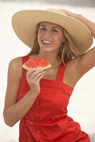 Junge blonde Frau im roten Sommerkleid und mit Sommerhut hält ein Stück Wassermelone