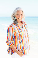 A mature woman with white hair on a beach wearing a striped shirt and white summer trousers