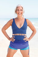 A mature woman with white hair on a beach wearing a blue bathing suit and turquoise shorts