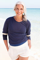 A mature woman with white hair on a beach wearing a blue top and white shorts