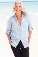 A mature woman with white hair on a beach wearing a light blue blouse and dark blue jeans