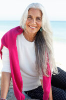A mature woman with white hair on a beach wearing a white jumper and polka dot trousers with a pink jumper over her shoulders