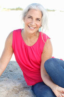 A mature woman with white hair on a beach wearing a pink top and blue jeans