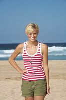 A blonde woman with short hair by the sea wearing shorts and a red-and-white striped top