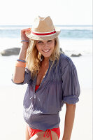 A blonde woman on a beach wearing a hat, a red bikini and a blue blouse