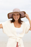 A brunette woman by the sea wearing a short-sleeved cardigan and a hat