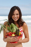 A young brunette woman on a beach wearing a bikini top and holding a bowl of vegetables