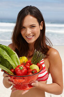 A young brunette woman on a beach wearing a bikini top and holding a bowl of vegetables