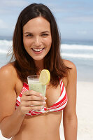 A young brunette woman on a beach with a smoothie wearing a bikini top