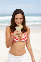A young brunette woman on a beach with a smoothie wearing a bikini top and white trousers