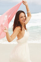 A brunette woman by the sea wearing a white summer dress and holding a pink shawl