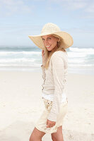 Blonde Frau mit Sommerhut in hellem Cardigan und Shorts am Strand