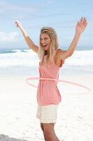 A young woman on a beach with a hula-hoop wearing a pink top and shorts