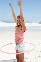 Junge Frau mit Hula-Hoop-Reifen im rosa Top und Jeansshorts am Strand