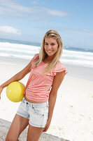 A young woman on a beach with a yellow ball wearing a pink top and denim shorts