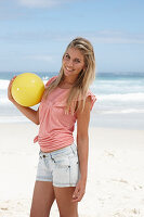 Junge Frau mit gelbem Ball im rosa Top und Jeansshorts am Strand