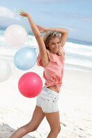 A young woman on a beach with balloons wearing a pink top and denim shorts