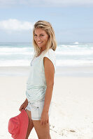 A blonde woman on the beach wearing a light t-shirt and denim shorts holding a red hat