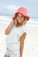 Blonde Frau mit rotem Hut in hellem T-Shirt und Jeansshorts am Strand