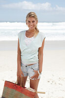 Blonde Frau mit Basttasche in hellem T-Shirt und Jeansshorts am Strand