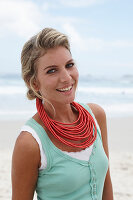 A blonde woman on a beach wearing a turquoise top and a red necklace