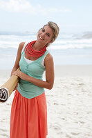 A blonde woman on a beach wearing a turquoise top, a red necklace and a salmon-pink skirt