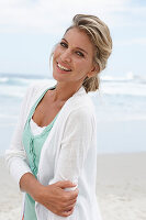 A blonde woman on a beach wearing a turquoise top and a white cardigan