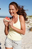 A brunette woman on a beach with a slice of watermelon wearing a light top and shorts