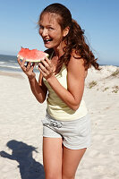 A brunette woman on a beach with a slice of watermelon wearing a light top and shorts