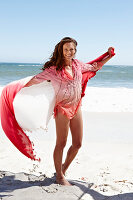 A brunette woman with a beach towel wearing a pink blouse and a bikini by the sea