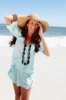 A brunette woman wearing a hat, a necklace and a light-blue beach dress by the sea