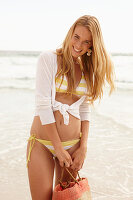 A blonde woman on a beach holding a bag wearing a white and yellow bikini and a cardigan