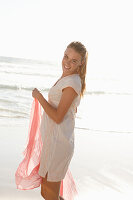 A blonde woman by the sea with a salmon-pink scarf and wearing a white dress