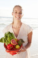 A blonde woman by the sea with a bowl of fruit and vegetables wearing a white dress