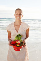 A blonde woman by the sea with a bowl of fruit and vegetables wearing a white dress
