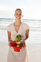 Blonde Frau mit Obst- und Gemüseschale in weißem Kleid am Meer